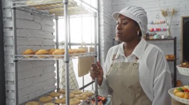 Pastanede önlük ve şapka takan genç bir kadın. Etrafı yeni pişmiş yiyeceklerle dolu raflarla çevrili. Arka planda çeşitli renkli pastalar ve tatlılar var.