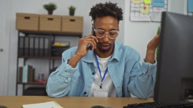 Ofiste çalışan, gözlük takan, telefonla konuşan, rafları ve bitkileri olan bir bilgisayar ekranına odaklanmış siyah bir adam..