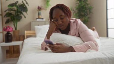 Genç bir kadın yatak odasında uzanıyor, telefona bakıyor, pembe pijamalar giyiyor, arka planda bitkiler var, rahatlama ve modern yaşam tarzını resmediyor, yetişkin bir kadın boş vakitlerden zevk alıyor..