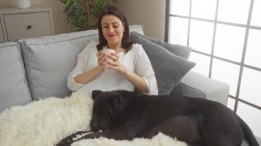 Latin kökenli genç bir kadın labrador köpeğiyle kanepede dinleniyor, rahat bir şehirde kahvesini yudumluyor..