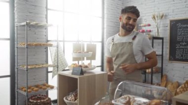 Ekmek, pasta ve kurabiyelerle çevrili bir fırında çalışan genç adam. Kapalı bir alanda kasayı kullanırken gülümsüyor.