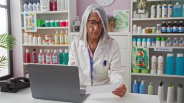 Bir eczanede çalışan yaşlı bir kadın, çeşitli ürünlerle çevrili, dizüstü bilgisayar kullanıyor.