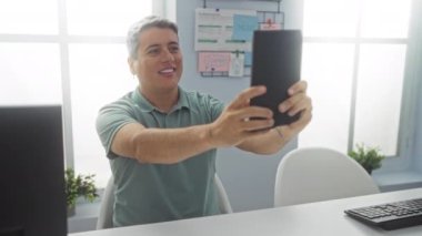 Ofisteki genç adam elinde tabletle selfie çekiyor. Etrafı ofis malzemeleri ve bitkilerle çevrili. Yeşil bir gömlek giyiyor..