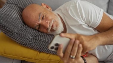 Sakallı ve gri saçlı orta yaşlı bir adam, gri bir yastığın üzerine uzanmış, rahat bir oturma odasında sarı bir yastık, beyaz bir gömlek giymiş, huzur içinde dinleniyor gibi görünüyor..