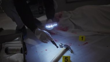 Adli tıp uzmanı, loş aydınlatılmış bir suç mahallinde kapalı alanda büyüteçle ve numaralandırılmış işaretlerle kanıtları inceliyor..