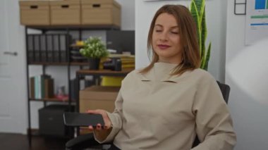 Genç, çekici, sarışın, beyaz bir kadın ofis ortamında, bir sandalyede oturuyor, telefonda konuşuyor, arka planda raflar, bitkiler ve ofis malzemeleri var..