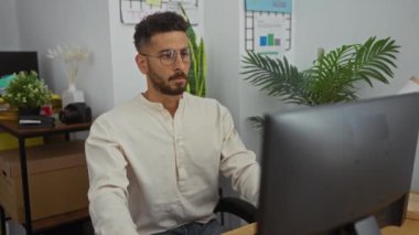 Genç İspanyol adam, bitki, kitap ve sabit eşyalarla dolu bir ofiste çalışırken kollarını uzatarak odaklanmış ve profesyonel bir iş ortamı yaratıyor..