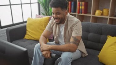 Gülümseyen siyah adam, modern dekorlu, aydınlık bir oturma odasında, gri bir koltukta öylesine oturuyor.