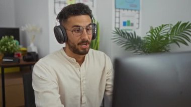 Ofis ortamında video konferans veren genç İspanyol bir adam, kulaklık ve gözlük takıyor, bitki ve çizelgelerle çevrili, iş aktiviteleriyle meşgul..