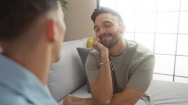 Bir adam, rahat bir apartman dairesinde birlikte otururken başka bir adama gülümsüyor. Modern bir ev ortamında, doğal ışık ve rahat bir atmosferde aşkı ve bağlantıyı resmediyor.