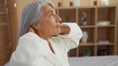 Gri saçlarını spa ortamında düzelten bir kadın, sağlık merkezinde orta yaşlı bir kadın, iç mekanda beyaz bir bornoz giyiyor, rahatlatıcı bir dekorla çevrili, rahatlatıcı bir atmosfer, olgun Kafkas güzelliği