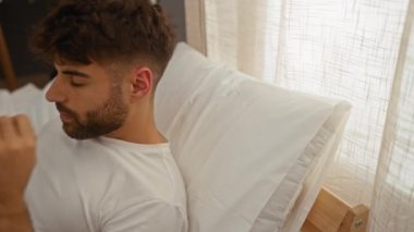 Genç adam yatak odasında uzanıyor, gözleri kapalı ve elleri kafasının arkasına sıkışmış bir şekilde dinleniyor, rahat ve sakin bir atmosferi evin içine taşıyor.