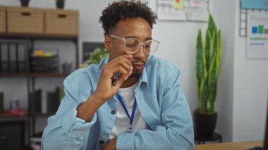 Gözlüklü genç Afro-Amerikalı adam modern bir ofis ortamında akıllı telefonuna dalgın dalgın bakıyor. Rafa kaldırılmış arka planı ve kapalı bitki dekoru var..