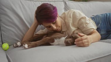 Mor saçlı genç bir kadın, rahat bir oturma odasındaki rahat bir kanepede uyuyan köpeğiyle sevgiyle yatıyor..