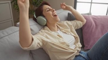 Kısa saçlı genç bir kadın evdeki koltukta kulaklıkla müzik dinliyor..
