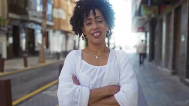 Kendine güvenen Afro-Amerikalı kadın şehir sokağında kollarını kavuşturmuş, doğal kıvırcık saçlarını ve dostça gülümsemesini sergiliyor..