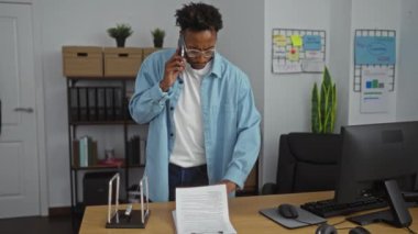 Mavi gömlekli Afro-Amerikalı genç bir adam, duvarında çizelgeler ve masasında belgeler olan modern bir ofiste masa başında çalışırken telefonuyla konuşuyor..