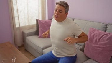 Evdeki oturma odasında rahat bir iç mekânla gerinen genç adam, gri kanepe ve pembe yastıklarla, kapalı alanda rahatlatıcı bir rutin sergiliyor..