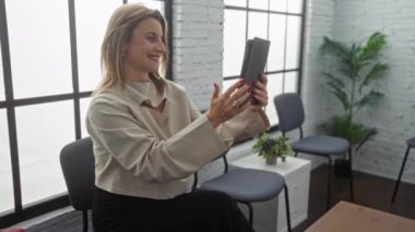 Sarışın, beyaz bir kadın modern kapalı bir bekleme odasında büyük bir pencerenin yanında sandalyeler ve bitkilerle birlikte bir tabletle selfie çekiyor..