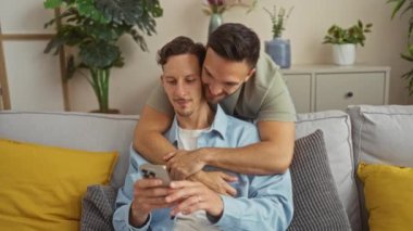 Sıcak oturma odasında kanepede kucaklaşan sevimli eşcinsel bir çift, bir adam telefonuna bakarken diğeri ona arkadan sarılıyor, yeşil bitkiler ve canlı yastıklarla çevrili.