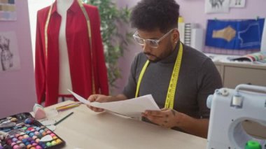 Odaklanmış bir adam canlı bir terzi dükkanında renkli kalemler ve dikiş makinesiyle moda tasarımlarını inceliyor..