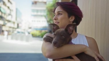 Gündüz vakti sokak ortasında köpeğini tutan genç bir kadın.