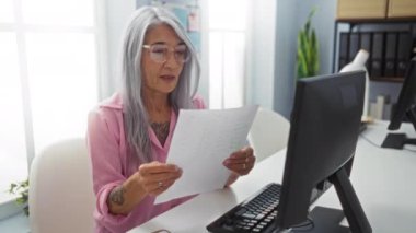 Ofiste bilgisayar başında çalışan, gözlük takan ve belge okuyan bir kadın mutlu ve profesyonel bir ortamda başarılı hisseder.