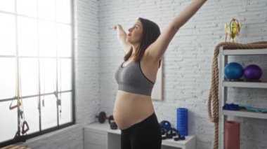 Hamile bir kadın spor salonunda egzersiz yapıyor. Etrafı spor malzemeleriyle çevrili yoga minderinin üzerinde geriniyor. Annelik egzersizlerini gösteriyor..