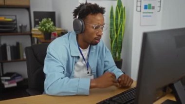 Sakallı ve gözlüklü Afrikalı genç bir Amerikalı ofisinde masasına oturmuş kulaklık takıyor ve mavi bir gömlek giyiyor. Bilgisayarıyla uğraşırken odaklanmış görünüyor..