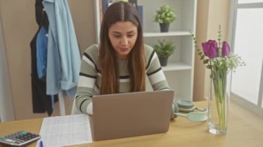 A young, focused hispanic woman works from home, celebrating her accomplishment on a laptop at a tidy living room table.