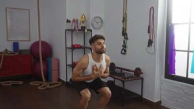 Spor salonunda spor yapan genç adam, çevresindeki ekipmanlarla spor yapıyor, atletik yapısını gösteriyor, beyaz tank giyiyor, ciddi ifade ediyor, çömelme pozisyonu, arka planda çeşitli ekipmanlar var.