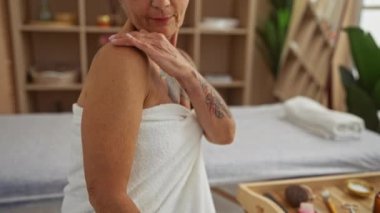 Beyaz havluyla omzuna losyon süren bir kadın sağlık ekipmanları ve yeşilliklerle çevrili bir spa ortamında kendini koruma ve rahatlama sergiliyor..