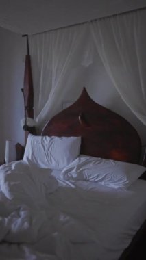 Loş ışıklı bir odada beyaz çarşaflı, eşsiz ahşap bir yatak başlığı ve katıksız perdeleri olan yapılmamış bir yatak..