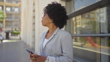Bir şehir caddesinde elinde telefon, gözlük ve gri bir ceket olan kıvırcık saçlı Afro-Amerikalı genç bir kadın..