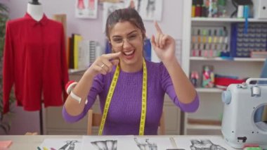Neşeli, genç İspanyol terzi tasarım odasının masasından seni işaret ediyor. Hareketli atölyesindeki kameraya gülümsüyor..