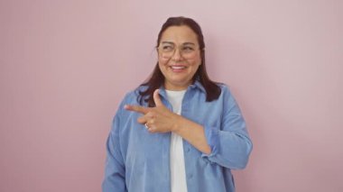 Neşeli orta yaşlı İspanyol kadın, gözlük takıyor, işaret ederken gülümsüyor, soyutlanmış pembe arka planda yan yana duruyor. Onun doğal, mutlu ifadesine hoş geldiniz.!