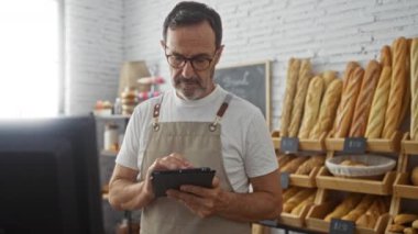 Fırında çalışan olgun bir adam ekmek dükkanında önlük giyerken tablet kullanıyor.
