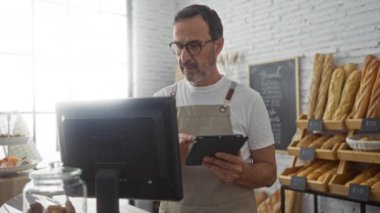 Fırında çalışan olgun bir İspanyol adam sergilenen taze ekmekle dolu bir odada tablet kullanıyor.