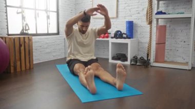 Genç, yakışıklı bir adam, beyaz tuğlalı duvarları olan, ağırlık, halat ve diğer spor aletleriyle çevrili, modern bir spor salonunda mavi bir paspasın üzerinde geriniyor..