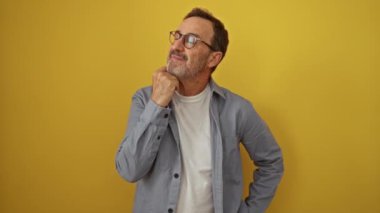 İspanyol orta yaşlı, gözlüklü, ayakta duran ve elleri çenesinde düşünen, izole edilmiş sarı bir arka plan, ciddi düşünceli bir yüz ve kafa karıştırıcı bir soru.