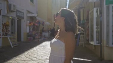 Güneş gözlüğü takan ve beyaz elbiseli genç bir İspanyol kadın, parlak güneş ışığı altında Gelibolu, Puglia, İtalya 'nın büyüleyici sokaklarını keşfediyor..