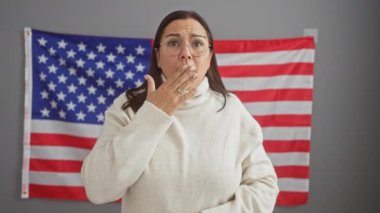 Şok olmuş orta yaşlı, gözlüklü İspanyol kadın bayraklı ofisinde dikiliyor, ağzını şaşkınlıkla kapatıyor, korkudan hata yapıyor.