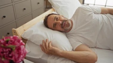 Bir yatak odasında yatarken kulaklarını kapatan İspanyol bir adam, muhtemelen gürültüden rahatsız olmuş, ön planda çiçekler olan bir ev ortamında.