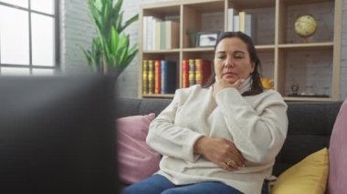 Düşüncelere dalmış olgun İspanyol bir kadın, evdeki kanepede oturuyor, sıradan bir zarafet sergiliyor..