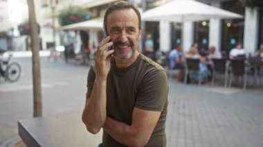Orta yaşlı bir adam, arka planda insanların oturduğu bir şehir merkezindeki bir kafede otururken cep telefonuyla konuşuyor.