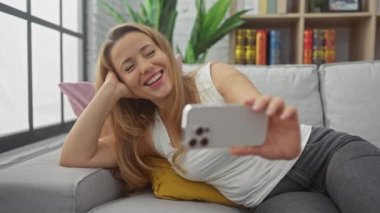 Sarışın kadın gülüyor ve evdeki kanepede akıllı telefonuyla selfie çekiyor..