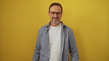 İspanyol orta yaşlı, gözlüklü, kolları çapraz duran, mutlu ve gülümseyen, izole edilmiş sarı bir arka plana karşı pozitif görünen bir adam.