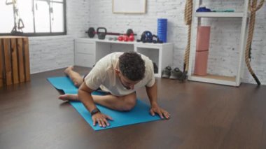 Arka planda çeşitli spor aletleri ve aksesuarlarla spor salonundaki minderde yoga yapan yakışıklı bir genç..