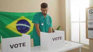 Tıbbi önlüklü genç bir İspanyol arka planda Brezilya bayrağı ve önünde iki oy kabini olan kapalı alanda oy kullanıyor..
