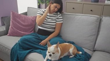 Çizgili tişörtlü genç bir kadın, gri bir kanepede Chihuahua köpeğiyle otururken, ev içinde rahat bir oturma odasında boyun ağrısı çekiyor..